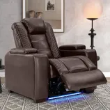 A brown recliner chair with quilted stitching is set in a cozy room, featuring a modern floor lamp, potted plant, framed artwork, and a small round side table with a speaker or device.