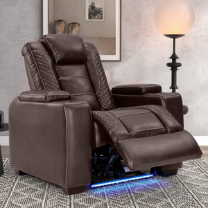 A brown recliner chair with quilted stitching is set in a cozy room, featuring a modern floor lamp, potted plant, framed artwork, and a small round side table with a speaker or device.