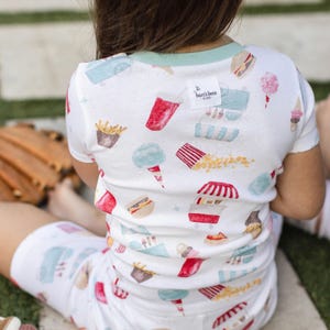 A child wears clothing with playful illustrations of fast food items like fries, popcorn, hot dogs, cotton candy, and soda. The label reads \“Burt's Bees Kids.\“