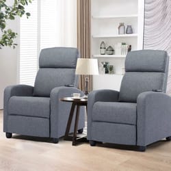 Set of two gray fabric massage recliners with cushioned high backs and armrests, positioned in a living room setting with a small round side table and lamp between them.