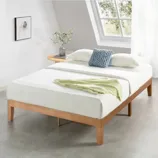 A minimalist wooden platform bed with a mattress, adorned with white sheets, a blue knitted pillow, and a light green throw. A round wooden nightstand holds a small plant and a book beneath an angled window.