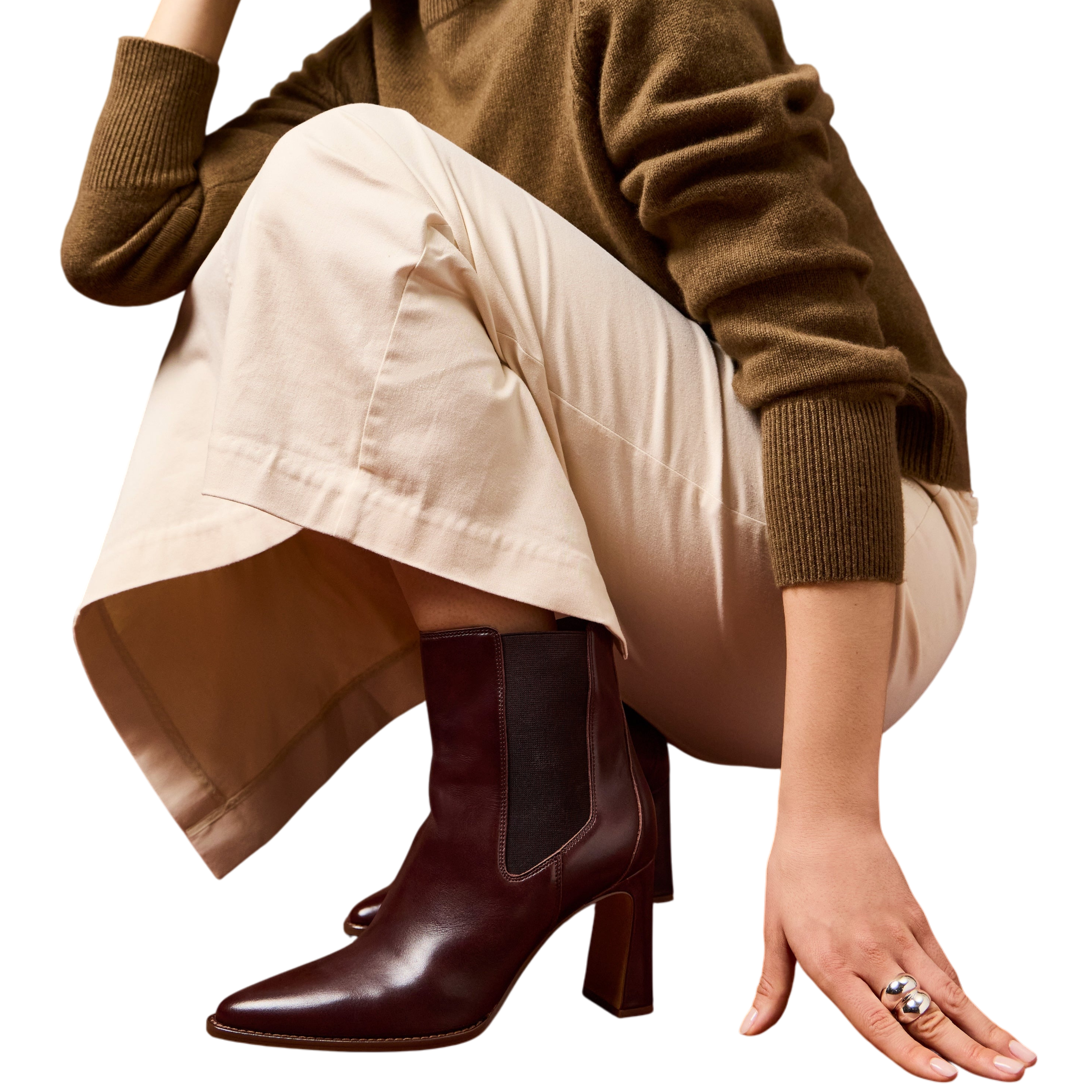 A person is shown wearing a brown sweater, cream pants, and heeled burgundy ankle boots with a dark elastic panel, alongside a silver ring with a dual orb design. The background is brown.