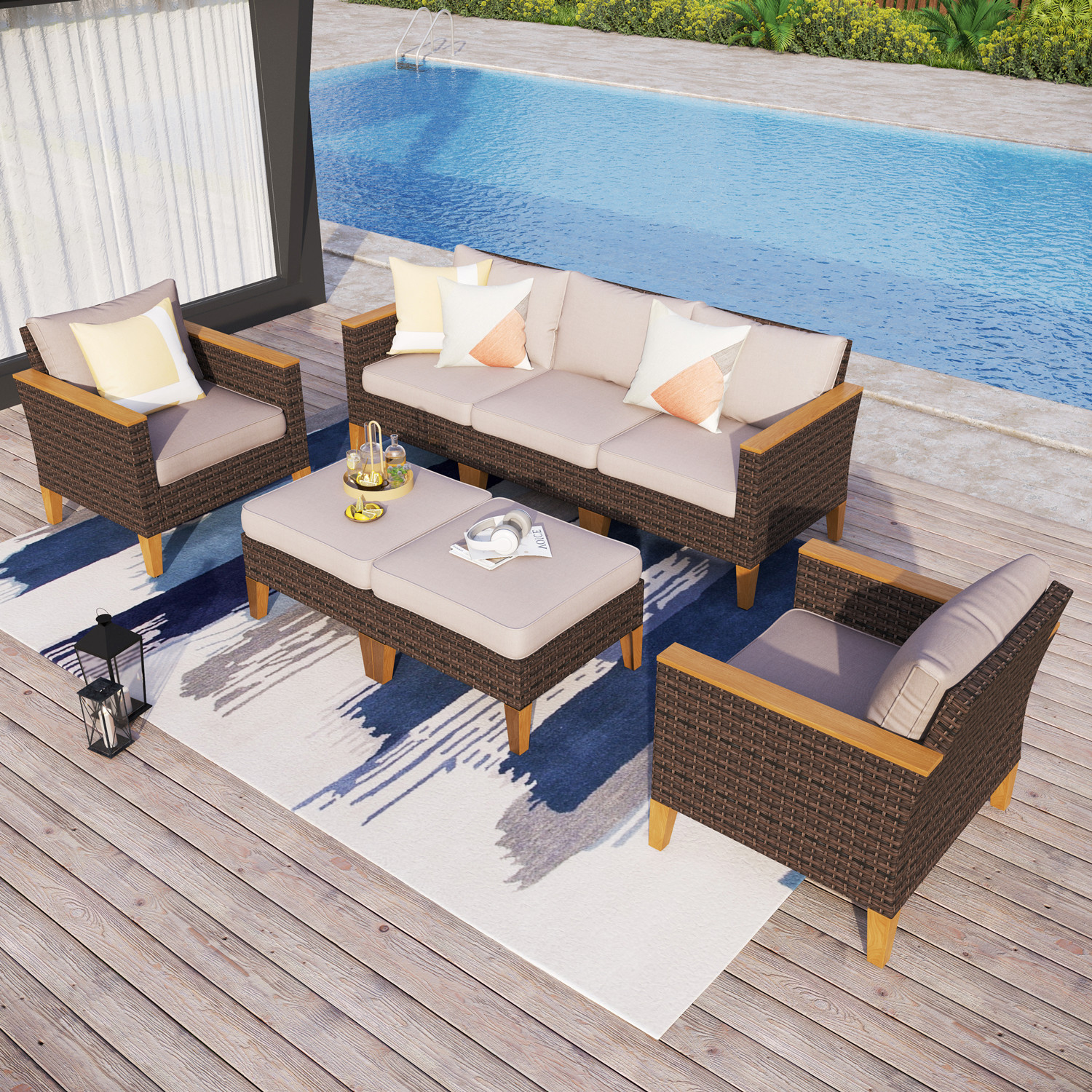 Outdoor seating set with a wicker sofa, two armchairs, and two ottomans, all featuring wooden legs and beige cushions. The setup includes a decorative rug, pillows, and a poolside view.