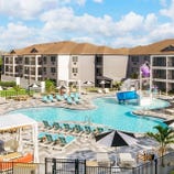 Outdoor resort pool area featuring lounge chairs, umbrellas, cabanas, and a children's water play area with a slide and splash bucket, set against a backdrop of multi-story hotel buildings.