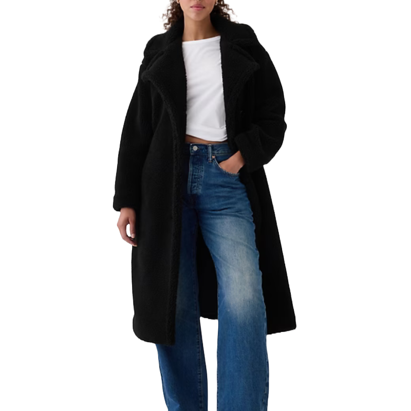 A woman is wearing a black Sherpa coat with a wide collar, long sleeves, and reaching mid-thigh, paired with a white t-shirt and blue jeans.