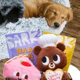 A dog sleeps in a fluffy bed next to BARK dog treats labeled as \“Baa Baa Jerky Bars.\“ Plush toys including a bear with a chef hat and a pink pastry with a strawberry are also visible.