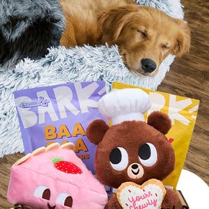 A dog sleeps in a fluffy bed next to BARK dog treats labeled as \“Baa Baa Jerky Bars.\“ Plush toys including a bear with a chef hat and a pink pastry with a strawberry are also visible.