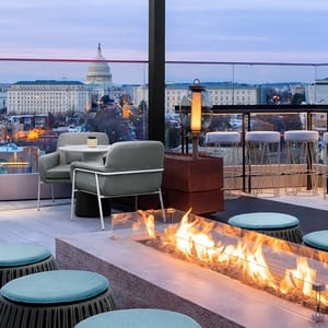 Rooftop patio with a fire feature, armchairs, and bar stools overlooking an urban landscape.