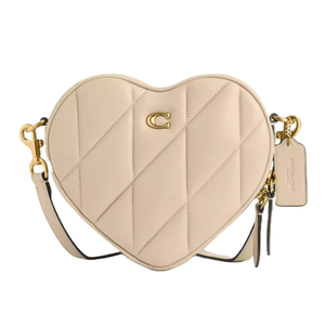 Heart-shaped Coach crossbody bag with quilted design, featuring a gold \“C\“ logo, detachable strap, and gold hardware in a soft beige color.
