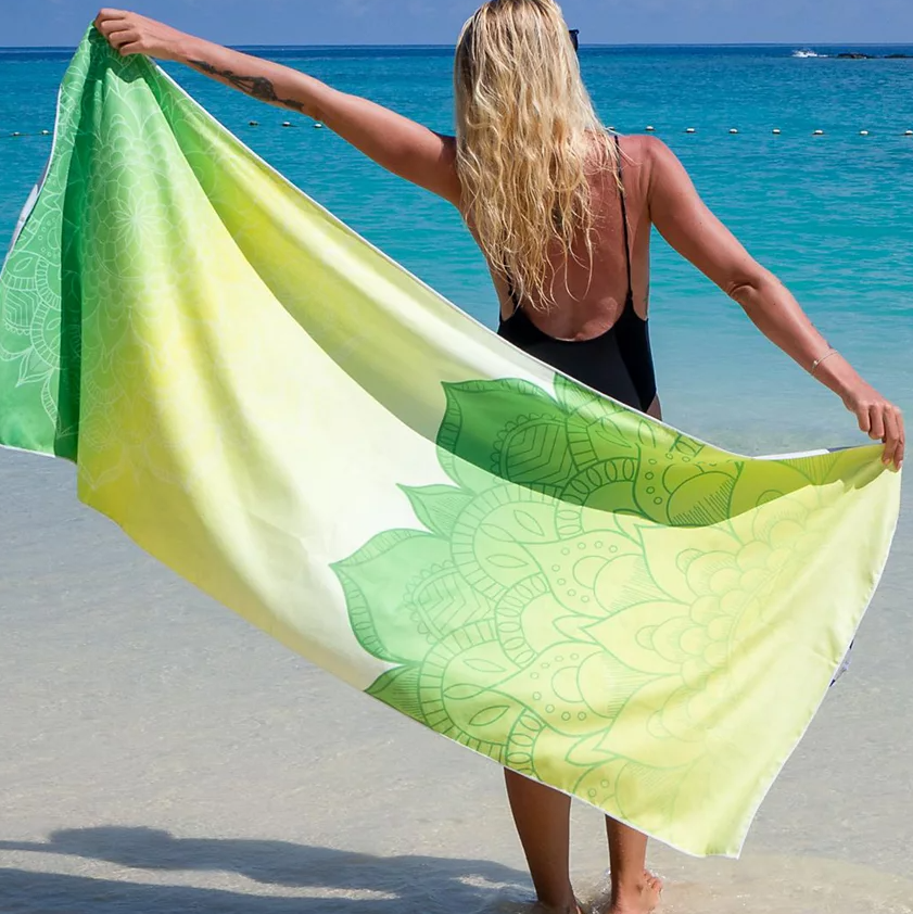 A person holds a large, 70-inch beach towel with a green and yellow mandala design, standing on a clear, sandy beach by turquoise water. The towel also comes with a travel bag.