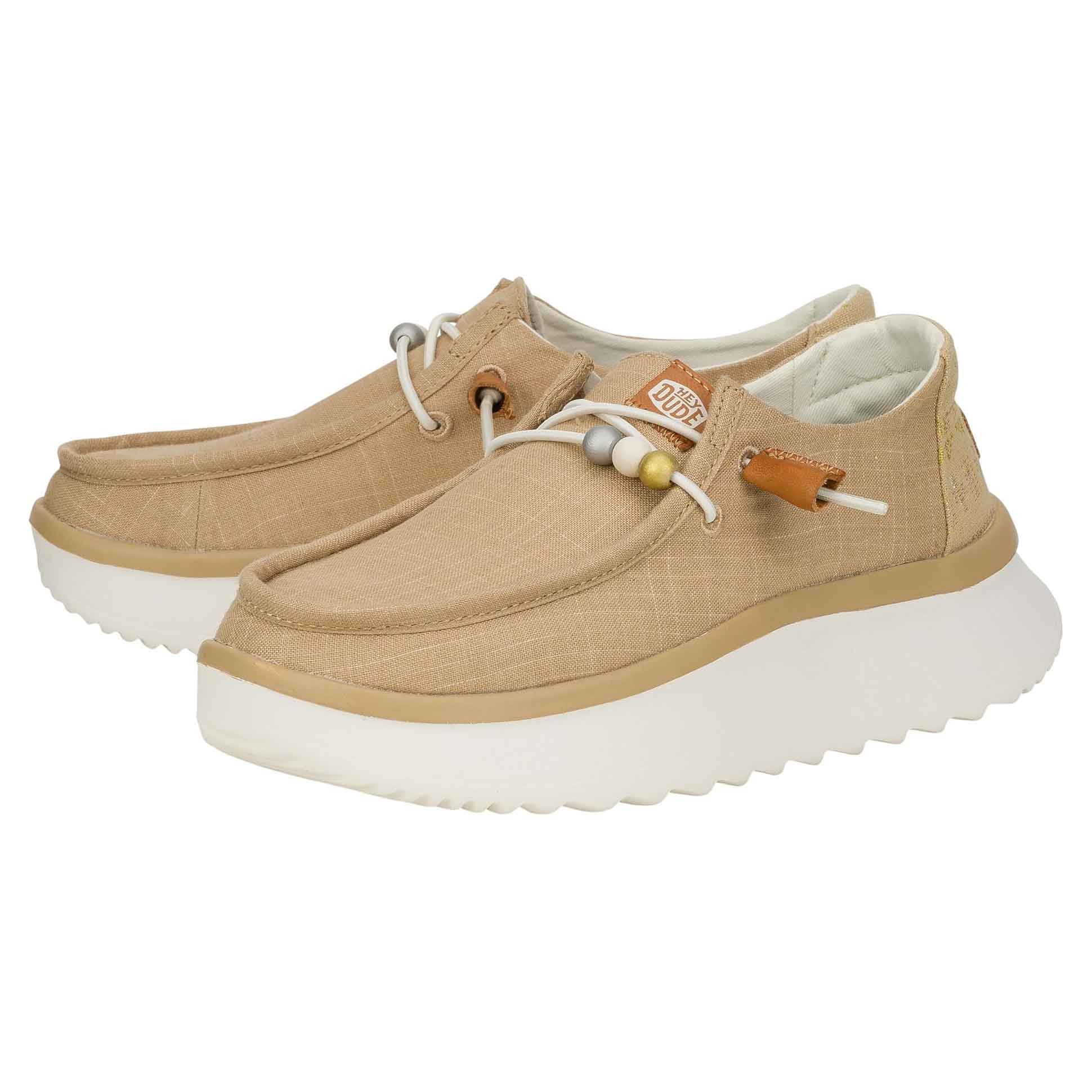 Hey Dude Wendy Peak Baja Stripe Shoes feature a beige fabric upper with a slip-on design, elastic laces, and decorative beads. They have thick white outsoles for comfortable wear.