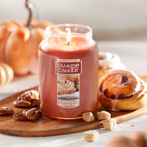 A lit Yankee Candle with \“Pumpkin Cinnamon Swirl\“ label sits on a wooden board beside cinnamon rolls and pecans.