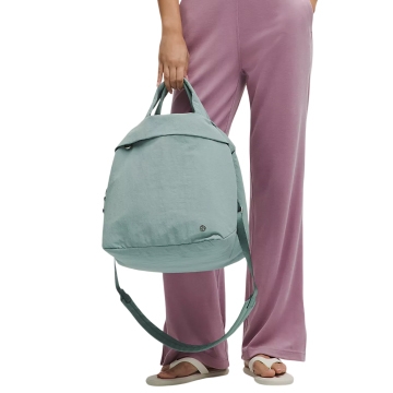 A person is holding a light blue lululemon 20L tote bag with dual handles and an adjustable shoulder strap, complemented by purple pants and white sandals.