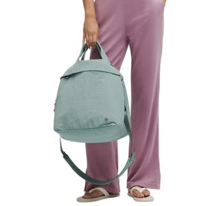 A person is holding a light blue lululemon 20L tote bag with dual handles and an adjustable shoulder strap, complemented by purple pants and white sandals.