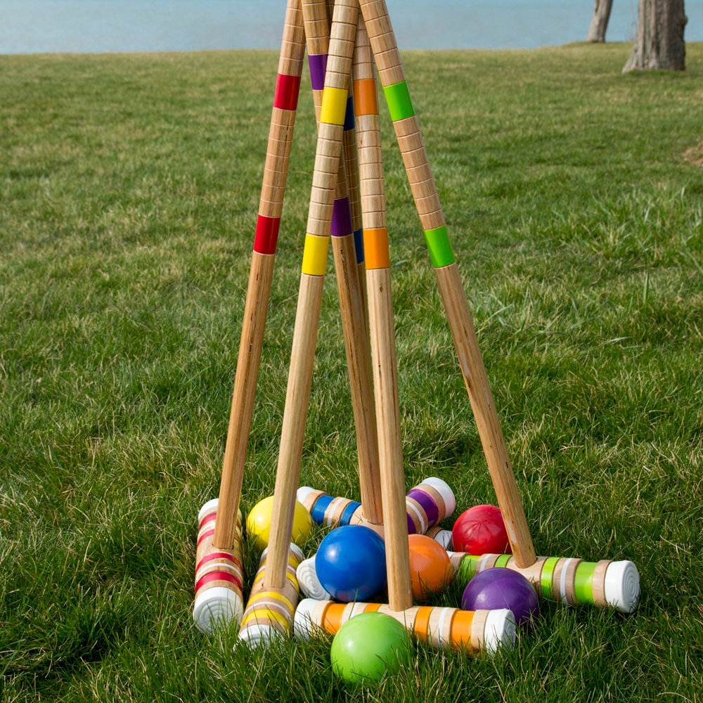 Six colorful wooden mallets with matching balls are laid on a grassy field, likely for a croquet game.