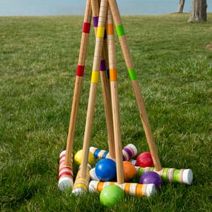 Six colorful wooden mallets with matching balls are laid on a grassy field, likely for a croquet game.