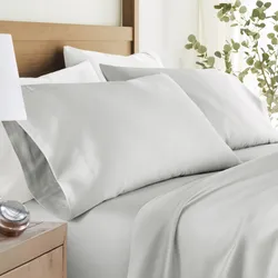 A neatly made bed with light gray pillowcases and bedsheets, alongside a wooden nightstand with a small clock. The bedding is styled in a calming, minimalist setting with green plant accents.