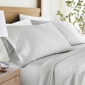 A neatly made bed with light gray pillowcases and bedsheets, alongside a wooden nightstand with a small clock. The bedding is styled in a calming, minimalist setting with green plant accents.