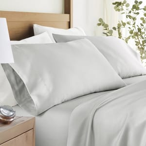 A neatly made bed with light gray pillowcases and bedsheets, alongside a wooden nightstand with a small clock. The bedding is styled in a calming, minimalist setting with green plant accents.