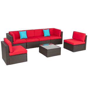 7-piece patio sectional set featuring a dark rattan frame with red cushions and two blue accent pillows includes a modular L-shaped sofa, two separate chairs, and a glass-topped coffee table, suitable for outdoor seating arrangements.
