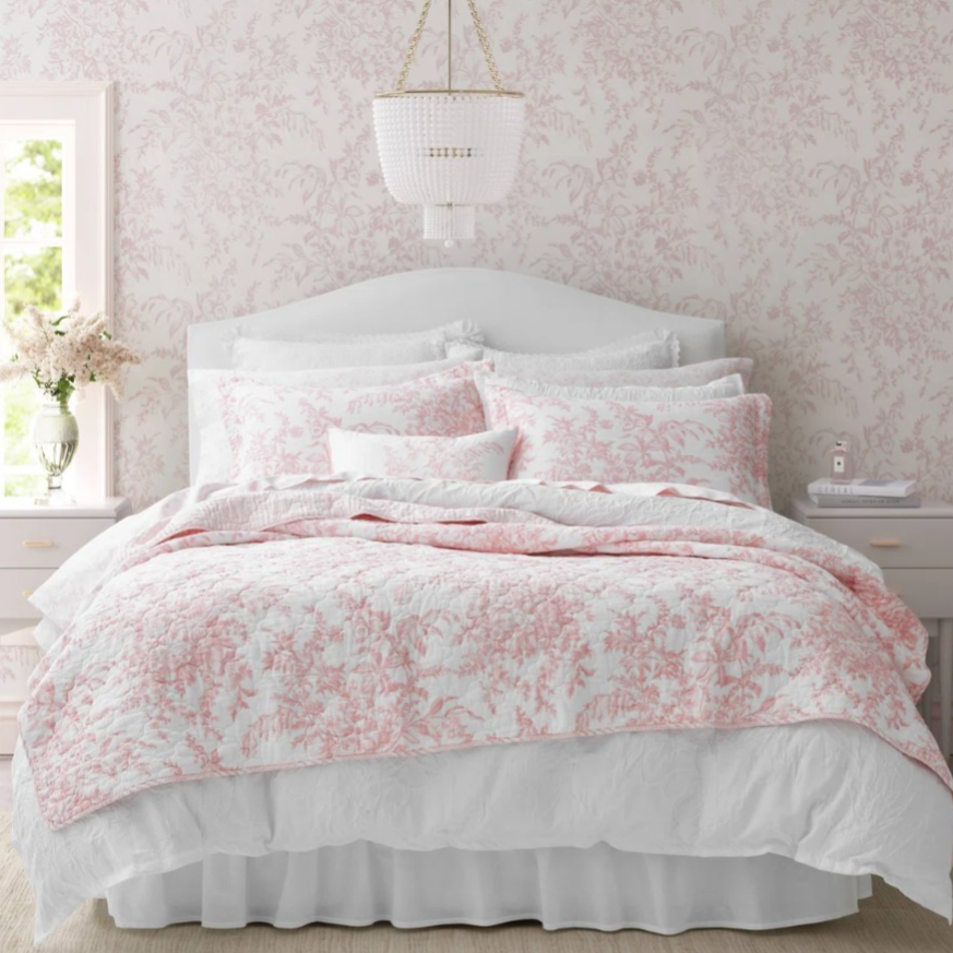 A bedroom features a white and pink floral-patterned comforter set on a bed with a white headboard. The wallpaper matches the bed's design, and a beaded chandelier hangs overhead. There are white bedside tables with a vase and a small decor item.