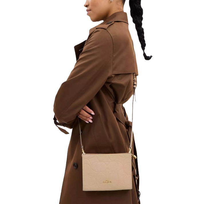 A person wears a brown trench coat carrying a beige crossbody bag embossed with a Coach logo.