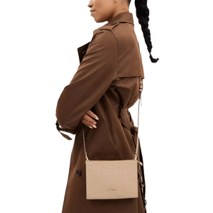 A person wears a brown trench coat carrying a beige crossbody bag embossed with a Coach logo.