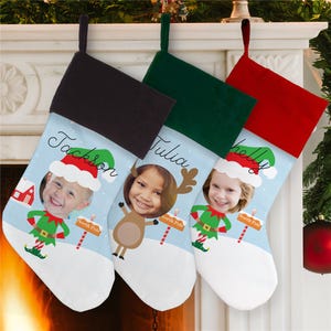 Three personalized Christmas stockings hang on a mantel, each featuring a child's face and name: Jackson, Tulia, and Shelly, with festive designs like elves and reindeer.