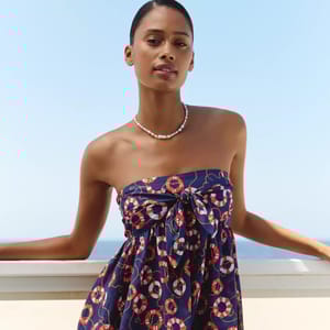 A woman is wearing a strapless dress with a nautical pattern and a pearl necklace. She stands against a railing with water and a blue sky in the background.