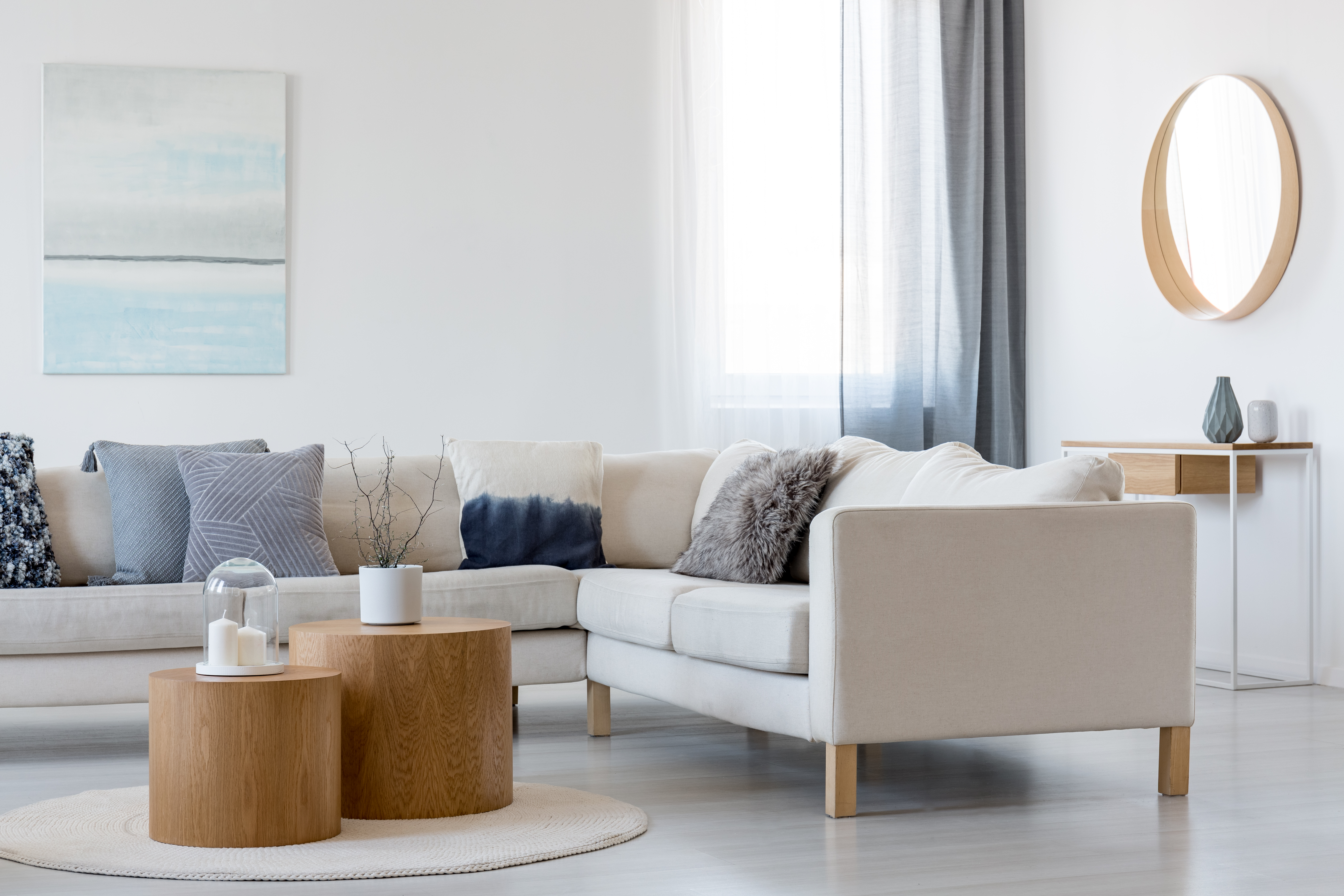 Modern living room setup with a beige sectional sofa, wooden nesting coffee tables, and decorative elements like pillows, a wall painting, and a round mirror.