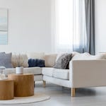 Modern living room setup with a beige sectional sofa, wooden nesting coffee tables, and decorative elements like pillows, a wall painting, and a round mirror.