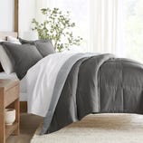 A bed is shown with a dark gray comforter and matching pillowcases on white sheets, next to a wooden nightstand and a potted plant.