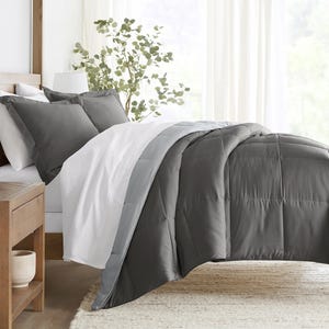 A bed is shown with a dark gray comforter and matching pillowcases on white sheets, next to a wooden nightstand and a potted plant.