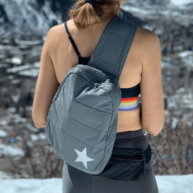 A person with a gray sling backpack featuring a white star, wearing a sports bra and a colorful headband. They're outdoors in a snowy mountain setting.