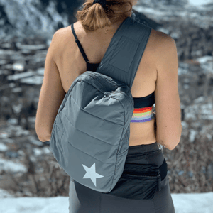 A person with a gray sling backpack featuring a white star, wearing a sports bra and a colorful headband. They're outdoors in a snowy mountain setting.