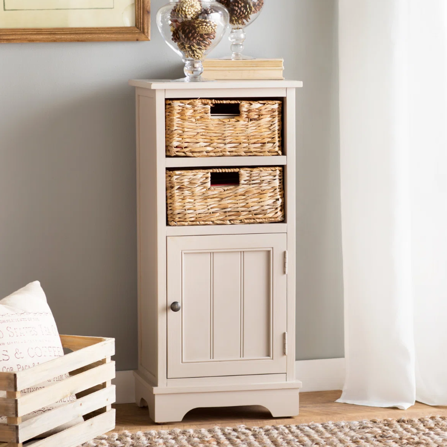 A beige cabinet with two woven baskets and a lower door sits next to a wooden crate and a neutral-toned rug, with framed artwork and decorative glass items on top.