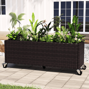 Rectangular wicker planter box on wheels, filled with various green plants and small white flowers, placed on a patio near a white building.