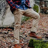 A person outdoors wearing khaki pants, brown leather boots, and a blue shirt with a red flannel jacket.