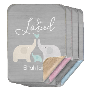 A soft gray Sherpa baby blanket featuring a graphic of three elephants with the words \“So Loved\“ and personalized with the name \“Elijah James\“ on the front. Several color layers are visible, including beige, pink, and blue.