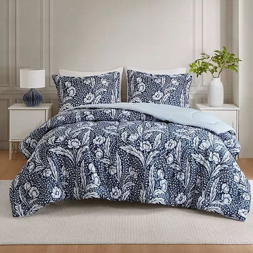 A bed is dressed with a navy blue and white floral patterned comforter set, including two matching pillow shams. The room features a white nightstand with a lamp and a vase with greenery.