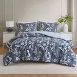 A bed is dressed with a navy blue and white floral patterned comforter set, including two matching pillow shams. The room features a white nightstand with a lamp and a vase with greenery.