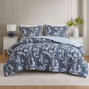 A bed is dressed with a navy blue and white floral patterned comforter set, including two matching pillow shams. The room features a white nightstand with a lamp and a vase with greenery.