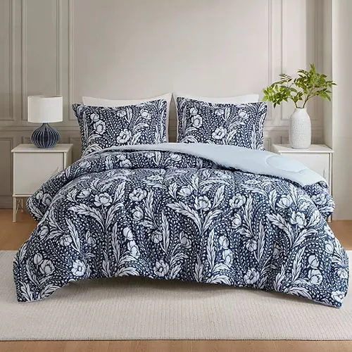 A bed is dressed with a navy blue and white floral patterned comforter set, including two matching pillow shams. The room features a white nightstand with a lamp and a vase with greenery.