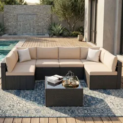 Outdoor patio set featuring beige cushioned sectional sofa with a dark frame, square coffee table with a reflective surface, blue patterned rug, and a decorative water feature and pool in the background.