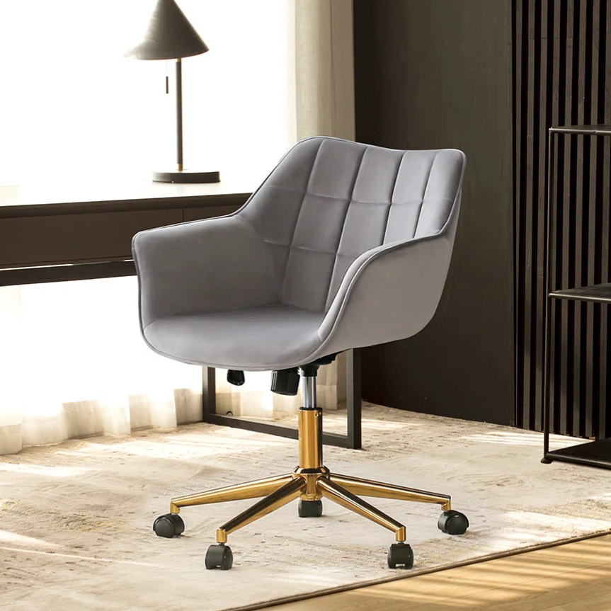 Gray swivel office chair with a quilted design and gold base on casters, placed in a modern office setting with a desk, lamp, and bookshelves.