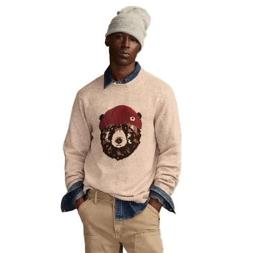 A man wears a light-colored sweater featuring a bear design, layered over a denim shirt. He's also wearing khaki pants and a gray beanie.