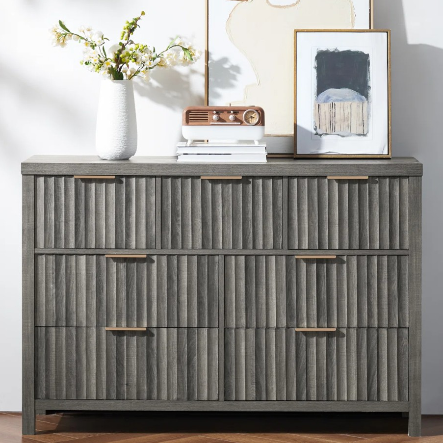 A gray dresser with vertical textured drawer fronts and brass handles, topped with a white vase with flowers, a small radio, and framed artwork.