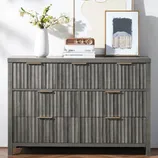 A gray dresser with vertical textured drawer fronts and brass handles, topped with a white vase with flowers, a small radio, and framed artwork.