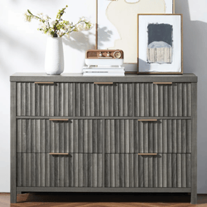 A gray dresser with vertical textured drawer fronts and brass handles, topped with a white vase with flowers, a small radio, and framed artwork.