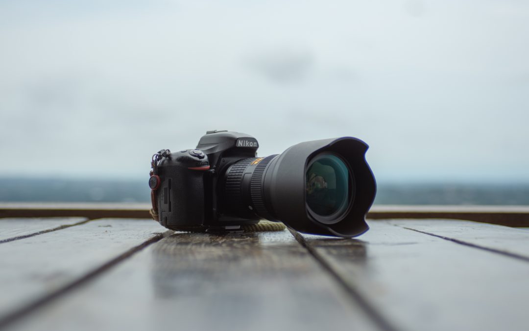 A DSLR camera with a telephoto lens is positioned on a wooden surface against a blurred waterscape background.
